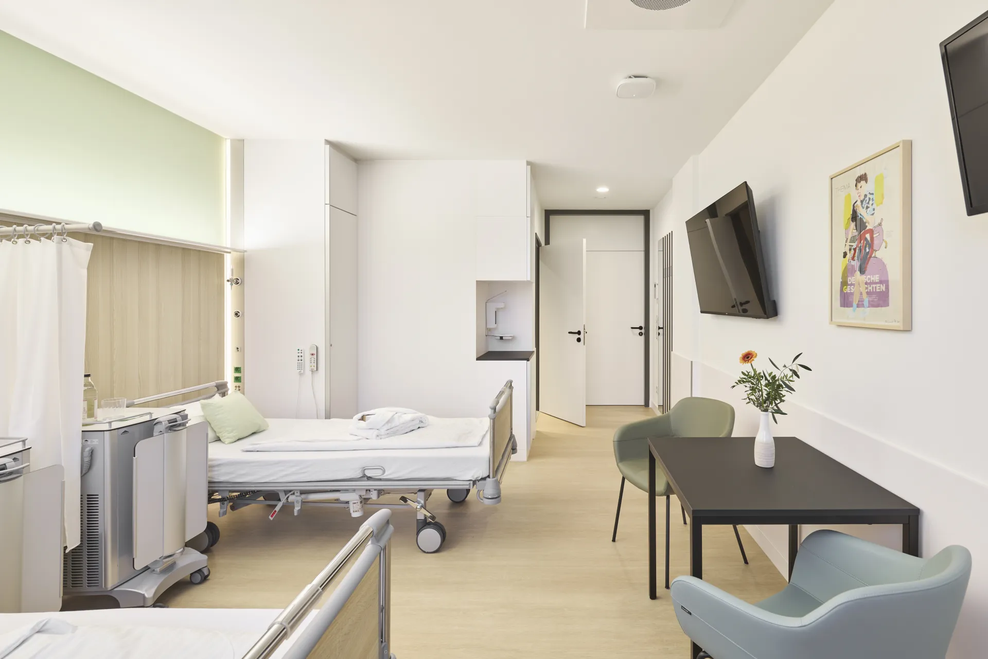 Helles Patientenzimmer mit zwei Betten, einem Tisch mit Stühlen, einem an der Wand montierten Fernseher und einem Blumenstrauß in einer Vase
